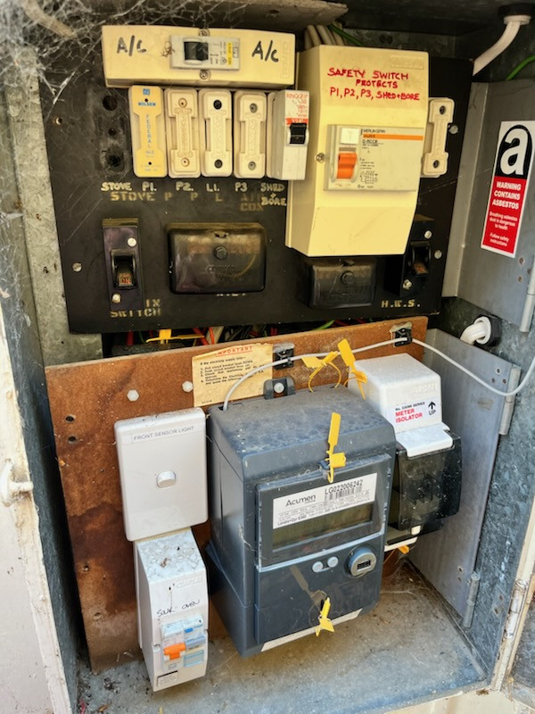 Real-world example of an old Australian switchboard showing asbestos warning sticker, ceramic rewirable fuses, corroded backing board, and no spare circuit breaker slots